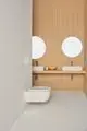 Minimalistische Badezimmereinrichtung mit Wandtoilette und zwei Waschbecken auf einem Holzregal. Ideal Standard Armatur und runde Spiegel über den Waschbecken.